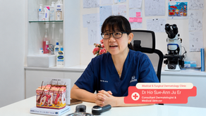 Dermatologist Dr Ho Sue-Ann Ju Er of Medical & Surgical Dermatology Clinic seated at her clinic desk next to a skin model anatomical display.