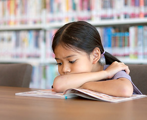 Young girl resting her head on a table with a tired expression, in a library or learning space, possibly showing signs of fatigue or illness, at Joyful Seeds Paediatrics located in Bukit Timah.