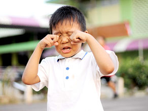 Young boy in a white shirt rubs his eyes, appearing upset. Blurred colorful background with greenery and a building. Joyful Seeds Paediatric and Developmental Clinic located in Bukit Timah
