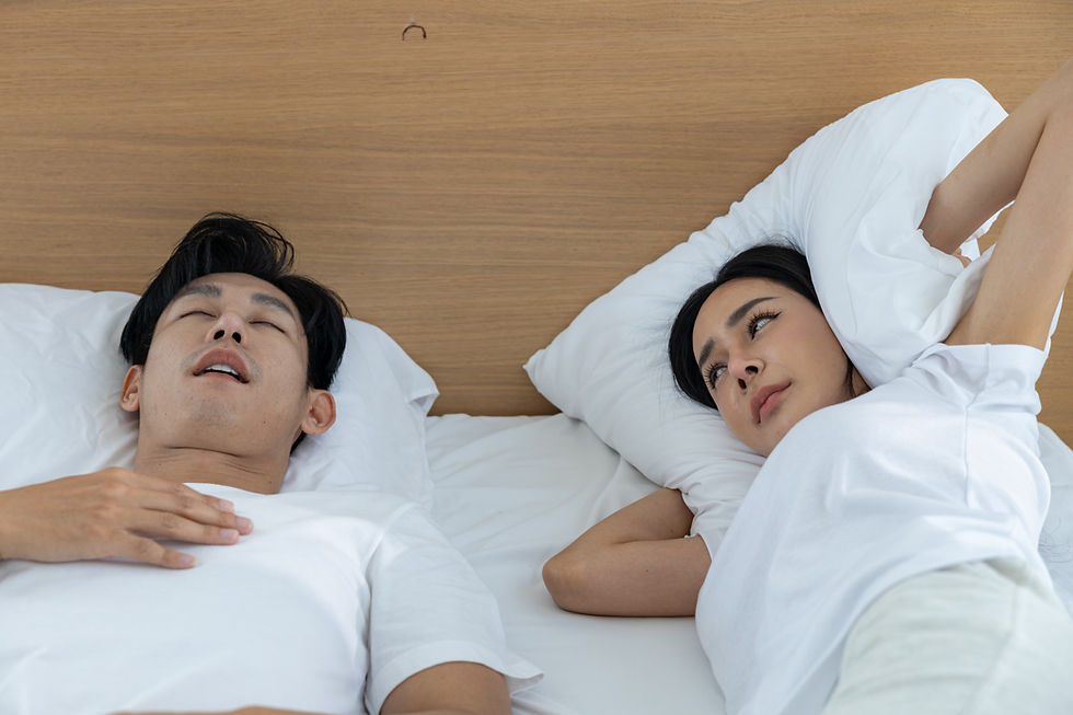 Couple lying in bed under white covers, man asleep snoring and woman awake.