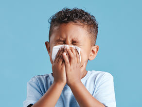 A child sneezes into a tissue, wearing a light blue shirt. Eyes closed, set against a solid blue background, conveying discomfort. Joyful Seeds Paediatric and Developmental Clinic located in Bukit Timah