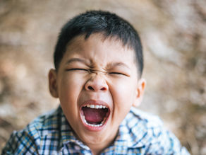 Young boy in a plaid shirt with eyes closed, mouth wide open, appearing to yell or shout, in an outdoor setting with blurred background. Joyful Seeds Paediatric and Developmental Clinic located in Bukit Timah