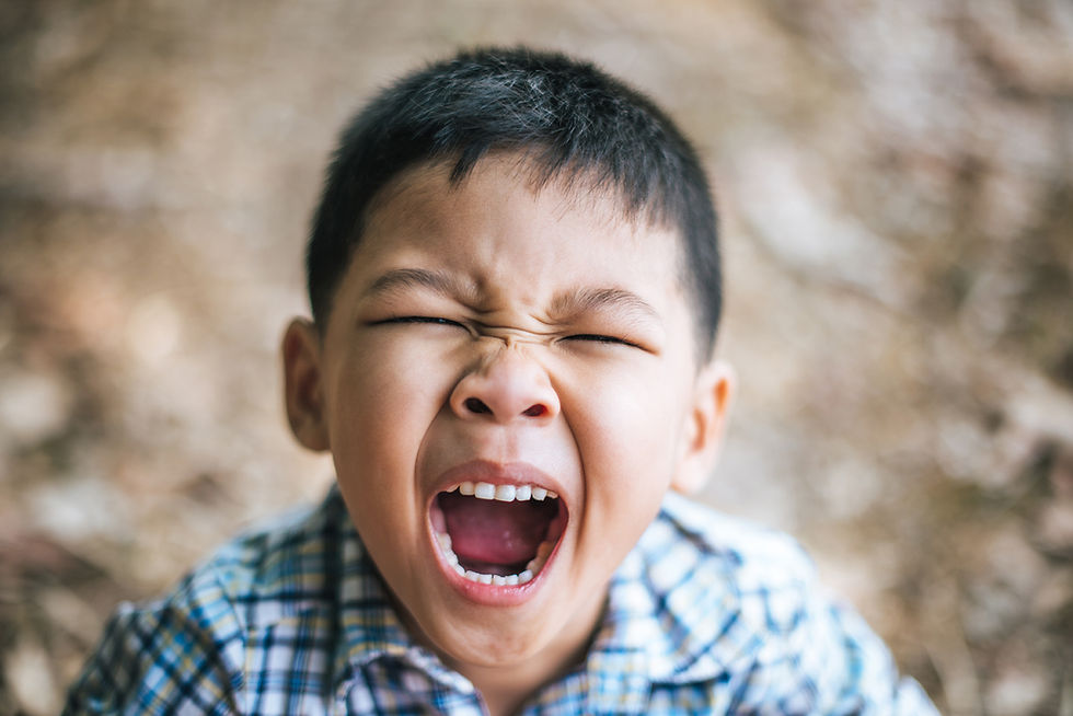 Young boy in a plaid shirt with eyes closed, mouth wide open, appearing to yell or shout, in an outdoor setting with blurred background. Joyful Seeds Paediatric and Developmental Clinic located in Bukit Timah