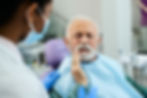 A dentist consults an elderly male patient who is touching his jaw, indicating tooth pain or discomfort, during a dental examination in a modern clinic setting