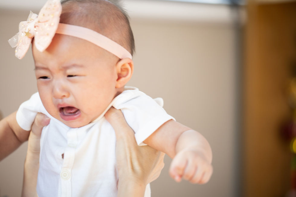 Crying baby in a white shirt and pink bow headband held by hands. Neutral indoor background, expressing distress.