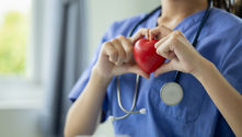 Person in blue scrubs and stethoscope holds a red heart shape between fingers. Soft light, calming atmosphere, blurred background.