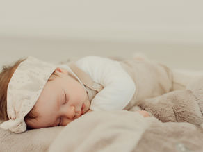 Sleeping baby in beige outfit and headband rests on a soft, fluffy surface. Neutral background evokes a sense of calm and coziness.