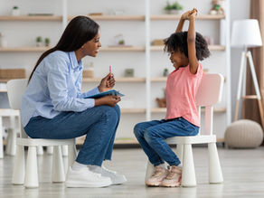 Woman and child in casual clothes sit on chairs in a bright room. The woman holds a pen, while the child raises their arms, smiling.