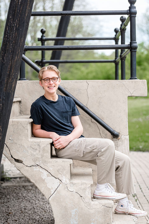 Batavia High school senior guy in a t-shirt and khaki pants sits on the steps leaning back and looking at the camera to his side mirroring the great angles of the steps of the Fabyan East Windmill.