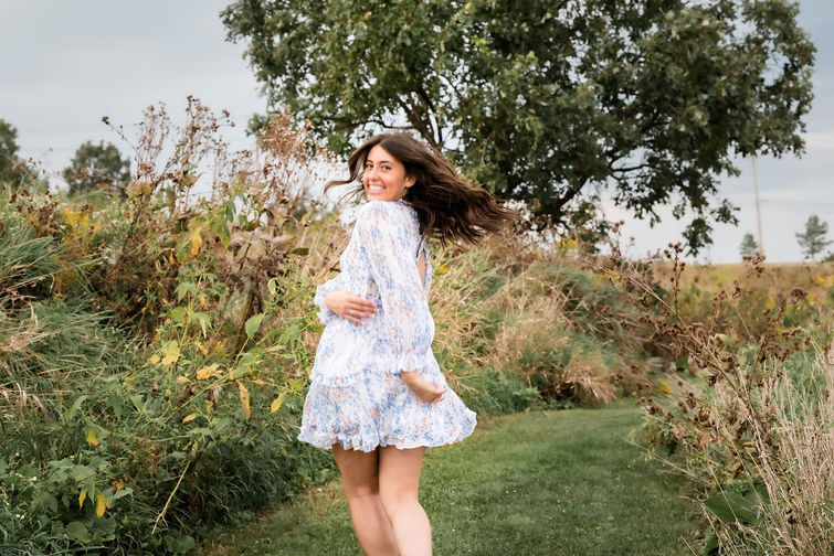 A Geneva senior girl looks over her shoulder and smiles as she playfully runs through a prairie path in a floral sundress with tall grasses and native flowers.
