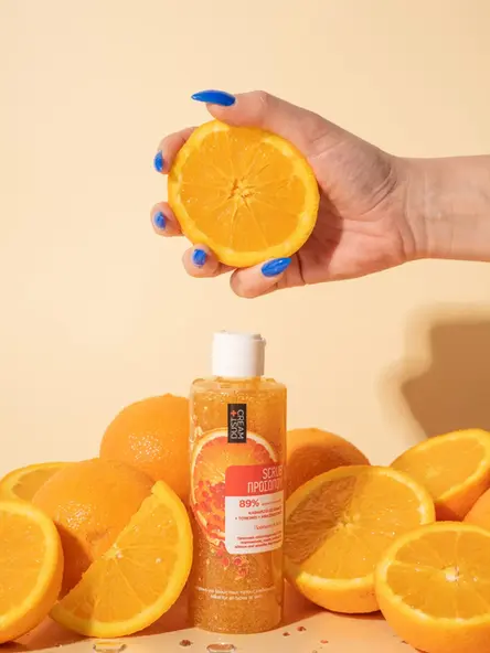 Scrub προσώπου Dust+Cream ανάμεσα σε φρέσκα πορτοκάλια και χέρι που κρατά μισό πορτοκάλι.