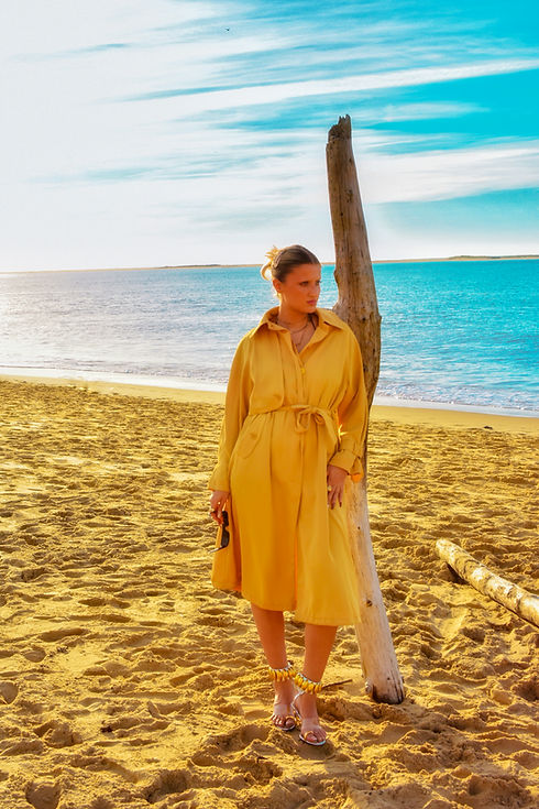 A person in a yellow dress stands on a sandy beach, leaning against a weathered wooden post. The ocean stretches out in the background under a clear blue sky with wispy clouds, as if captured by an artiste photographes lens.