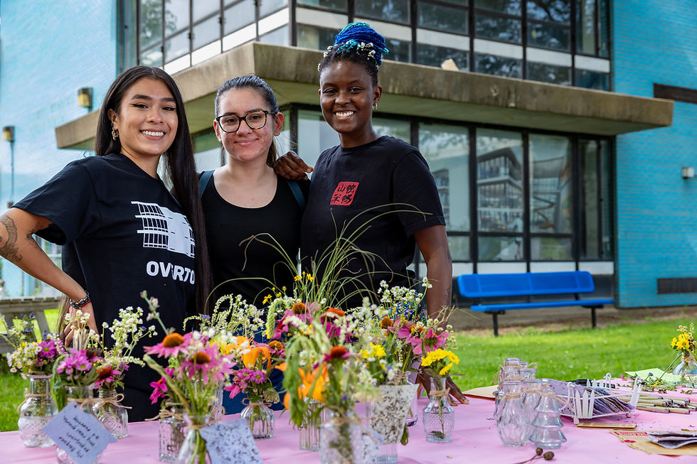 Jenny Guzman and two attendees smile in front of the Overton building. Just in front of them is a table with many small, colorful bouquets.