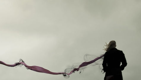 Woman stands on a grassy hill in windy gray sky, black coat and purple scarf streaming behind her in a moody scene