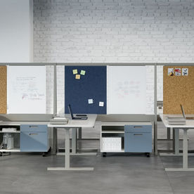 Modern workstation featuring individual desks with adjustable heights, blue and white storage units, and partition panels with corkboards, whiteboards, and fabric pinboards. The workspace is set against a white brick wall, offering an open and collaborative environment.