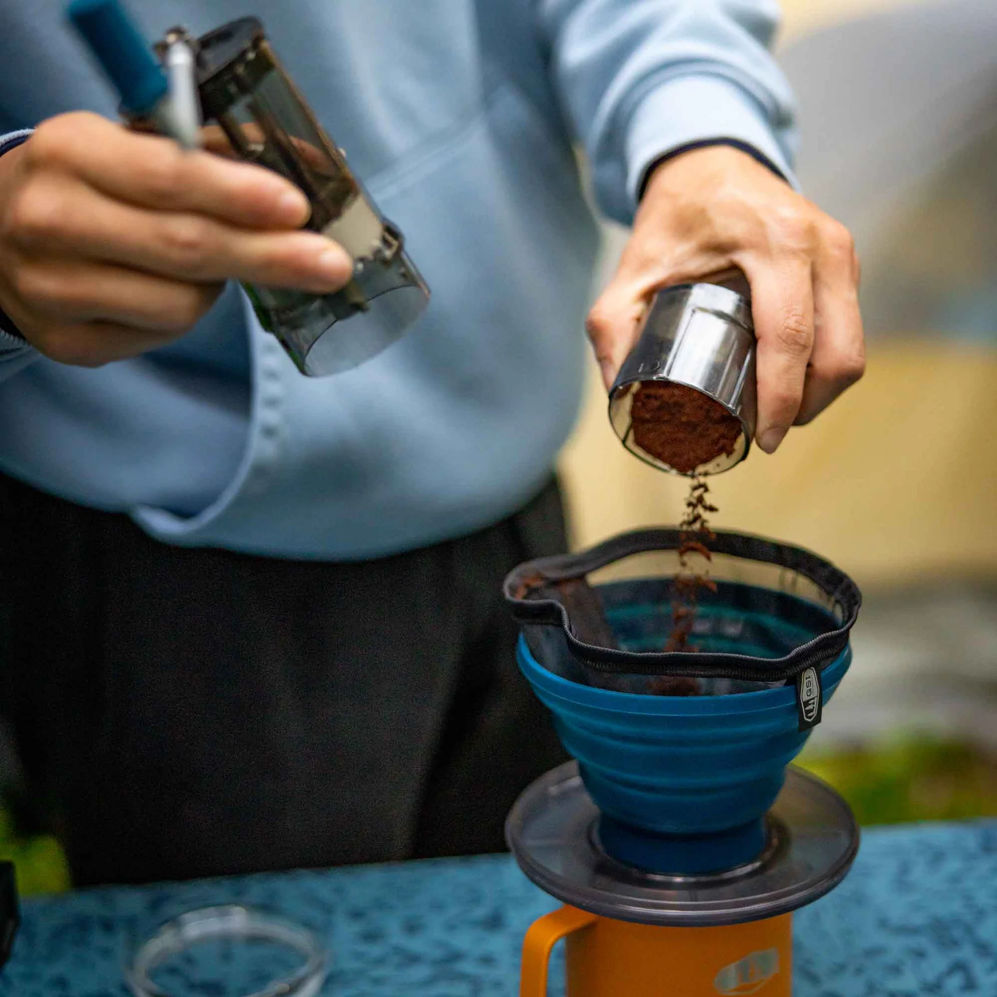 Person pouring ground coffee into the GSI Outdoors Collapsible Java Drip coffee maker while preparing a fresh brew outdoors.