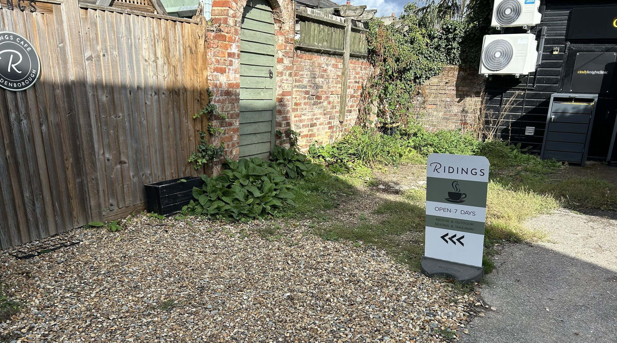 Sunlit back courtyard of rear gate entrance of Ridings Café in Hildenborough by brick wall and wooden fence, with a Ridings cafe sign: Open 7 Days, indoor & outdoor seating and takeaway.