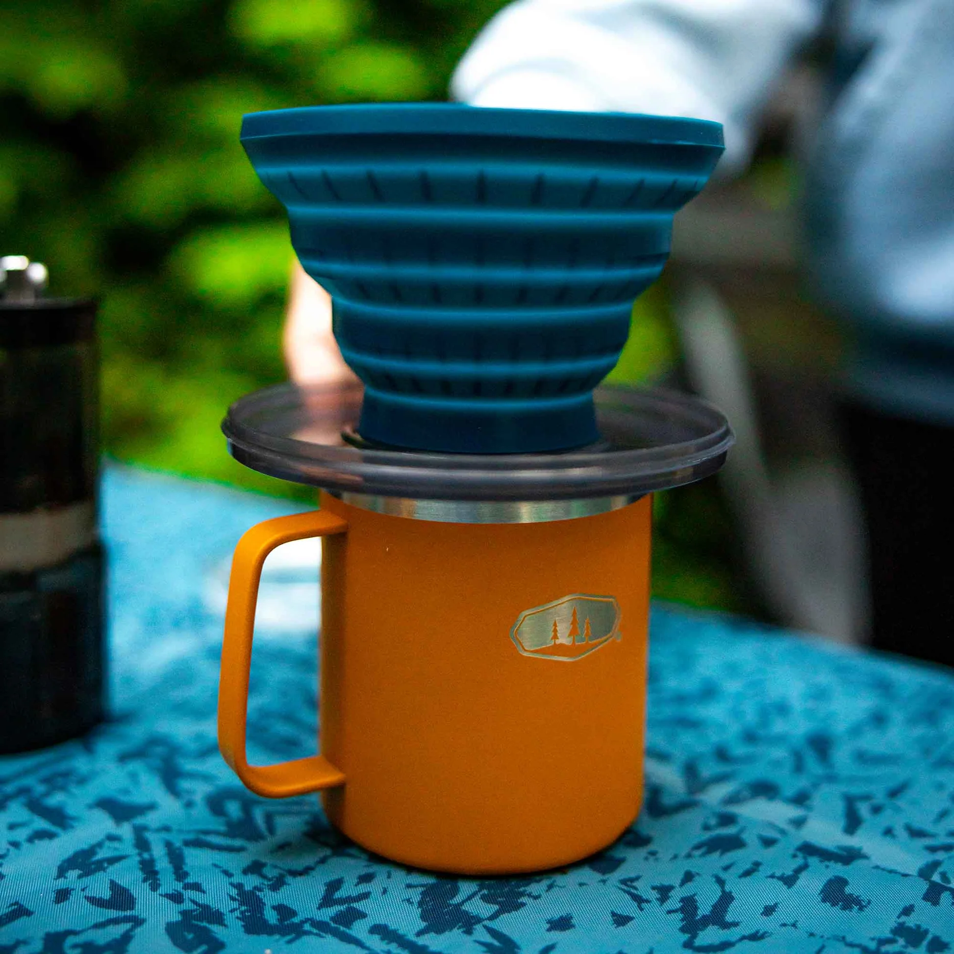 Close-up of the GSI Outdoors Collapsible Java Drip coffee maker set on an orange camping mug on a patterned blue tablecloth.