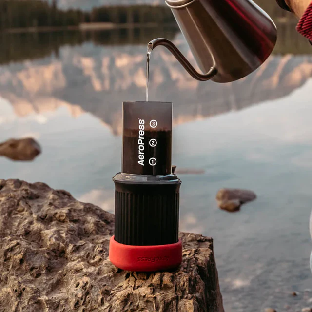A person pouring hot water into an AeroPress Go coffee maker on a rock by a scenic lake, with mountains reflecting in the water.