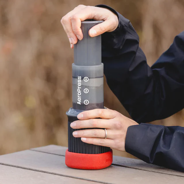 Close-up of hands pressing down on an AeroPress Go coffee maker while brewing coffee outdoors on a wooden surface.