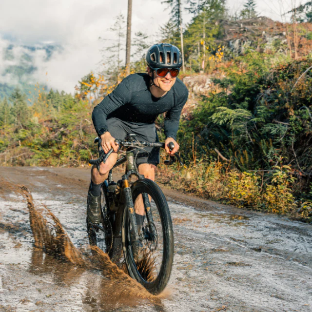 A cyclist riding a gravel bike through a puddle wearing the Vallon Watchtowers sunglasses