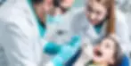 Dentists in white coats and gloves examine a child's teeth in a dental office. The child looks cheerful. Bright lighting and clean setting.
