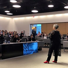 A speaker in red shoes addresses an audience in a modern conference room, with attendees engaged and laptops open.