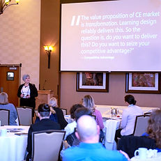 A speaker presents in front of an audience, with a quote about transformation displayed on a screen behind them.