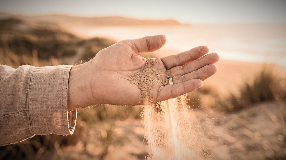 Close-up of a sandy hand letting sand trickle down at a sunlit beach, with warm blurred coastline in the background.