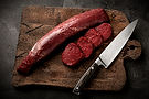 Premium cuts of African game venison meat including a dried sausage and sliced fillets with a butcher knife on a dark wooden board