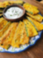 Golden breaded zucchini slices encircle a wooden bowl of creamy dip on a floral-patterned plate, set on a rustic wooden table.