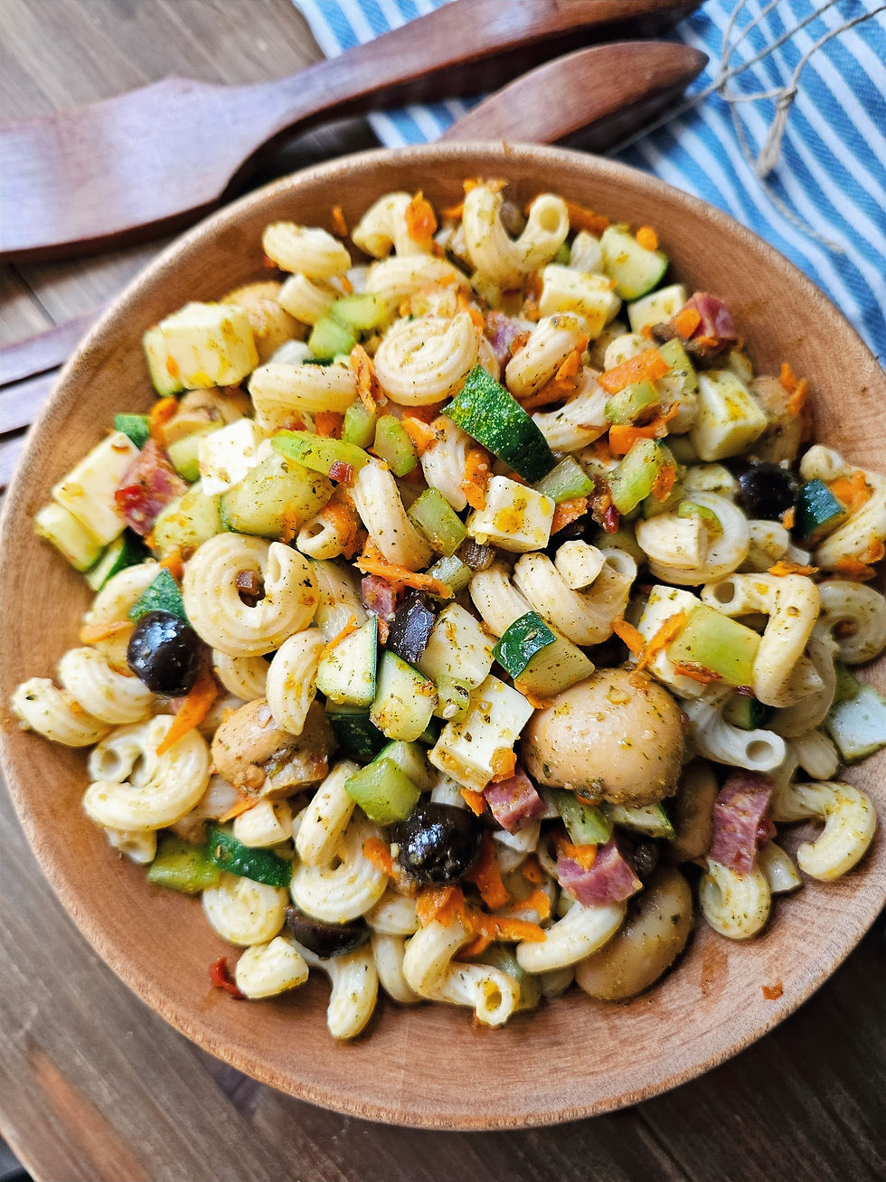 Bowl of pasta salad with zucchini, carrots, olives, and salami on a wooden table. Striped cloth and wooden utensils in the background.