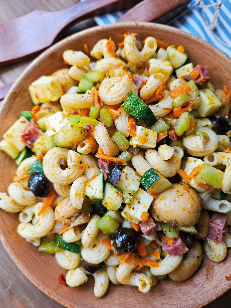A wooden bowl of pasta salad with colorful veggies, olives, and cheese on a wooden table, with a striped cloth in the background.