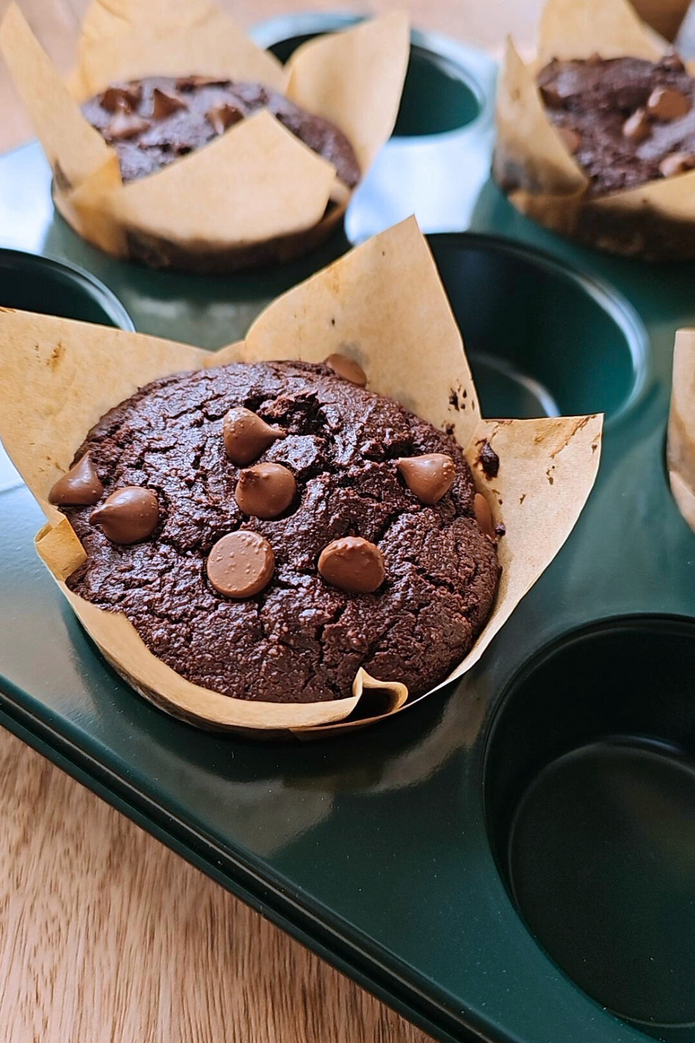 Chocolate muffins with chocolate chips in brown parchment, placed in a dark green muffin tin on a wooden table. Warm and inviting.