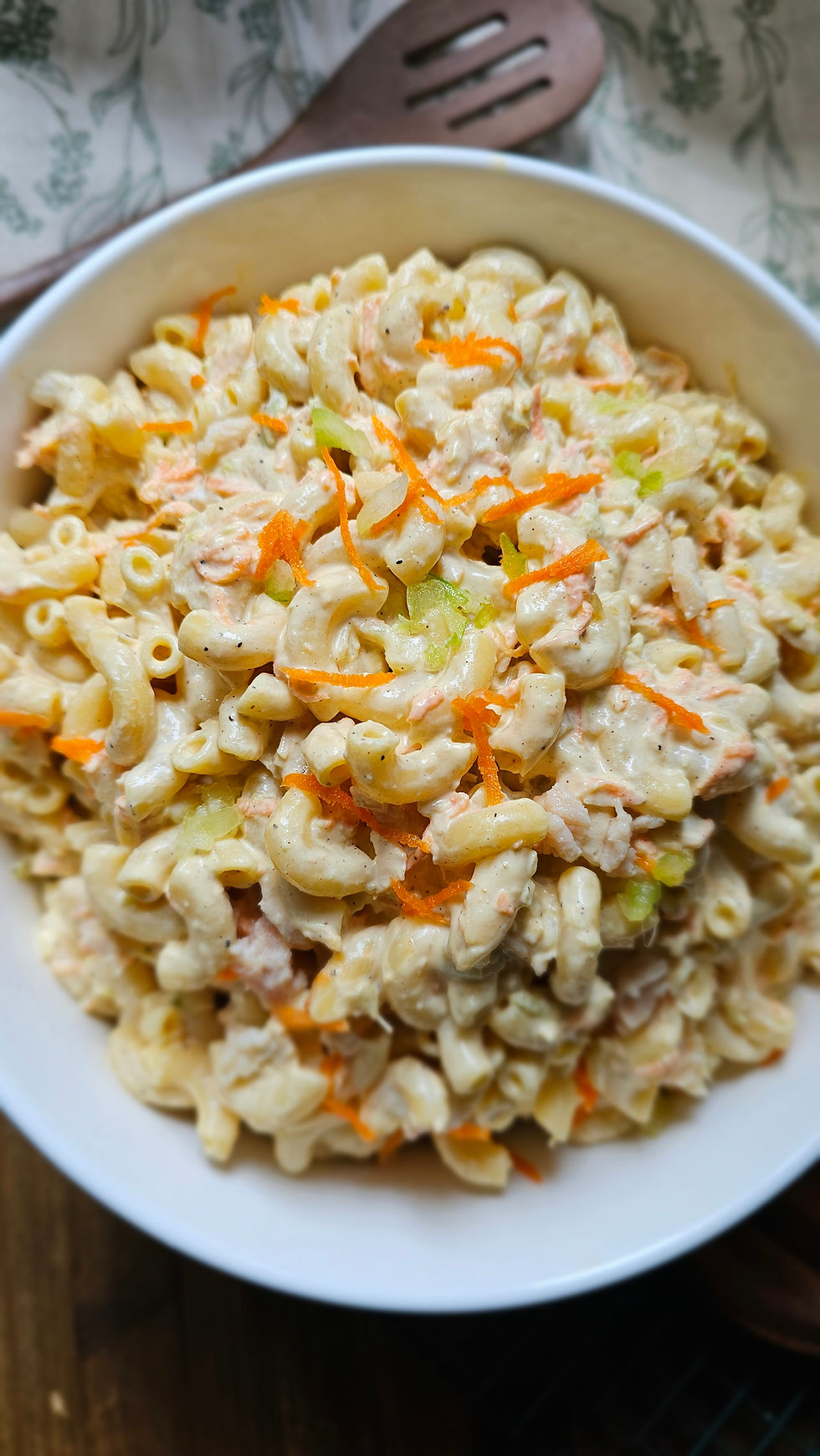 Bowl of creamy macaroni salad topped with shredded carrot and celery, with a wooden spoon on a patterned table.