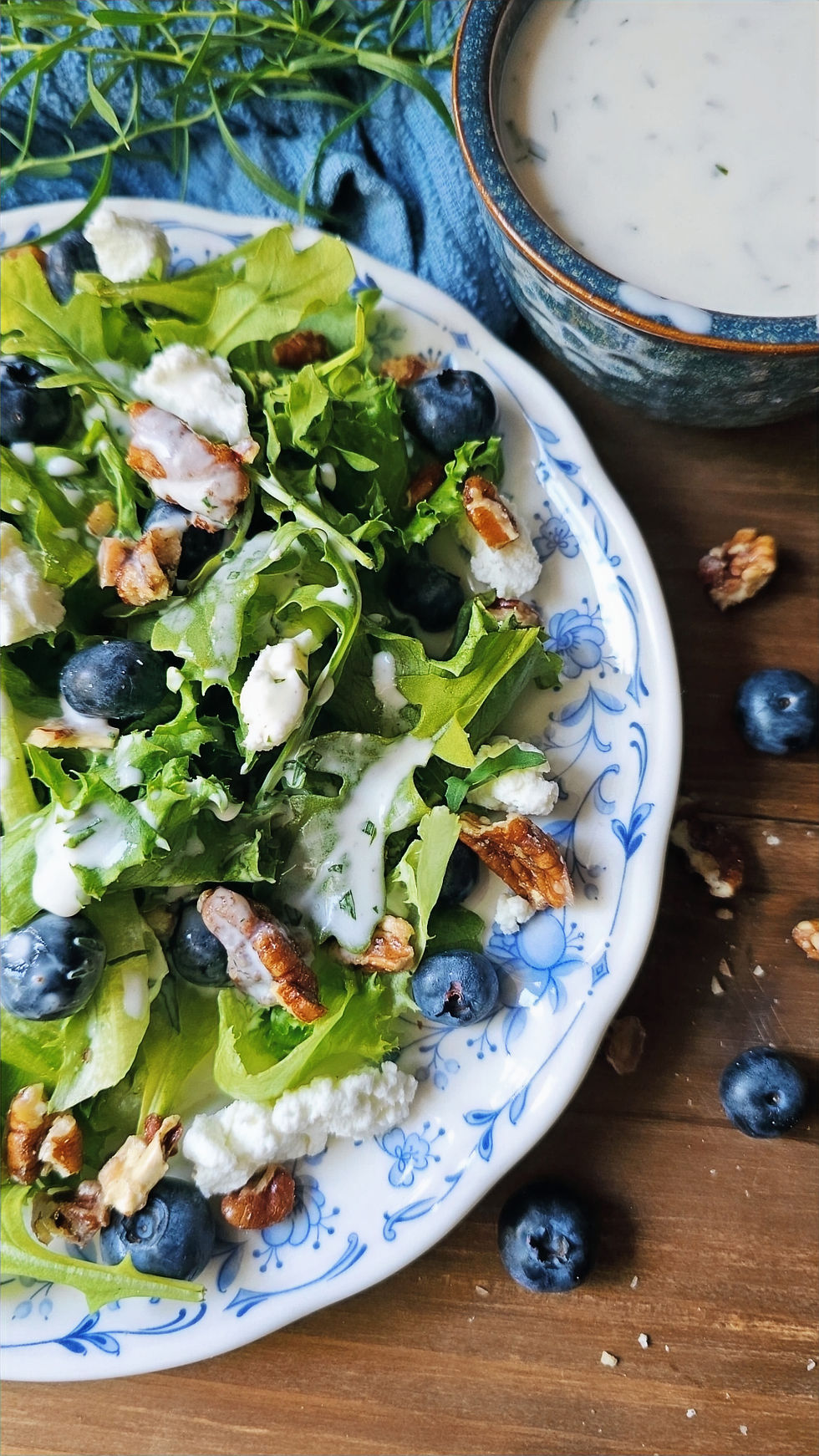Fresh green salad with blueberries, pecans, and crumbled goat cheese on a blue floral plate beside a bowl of creamy soup on wood table