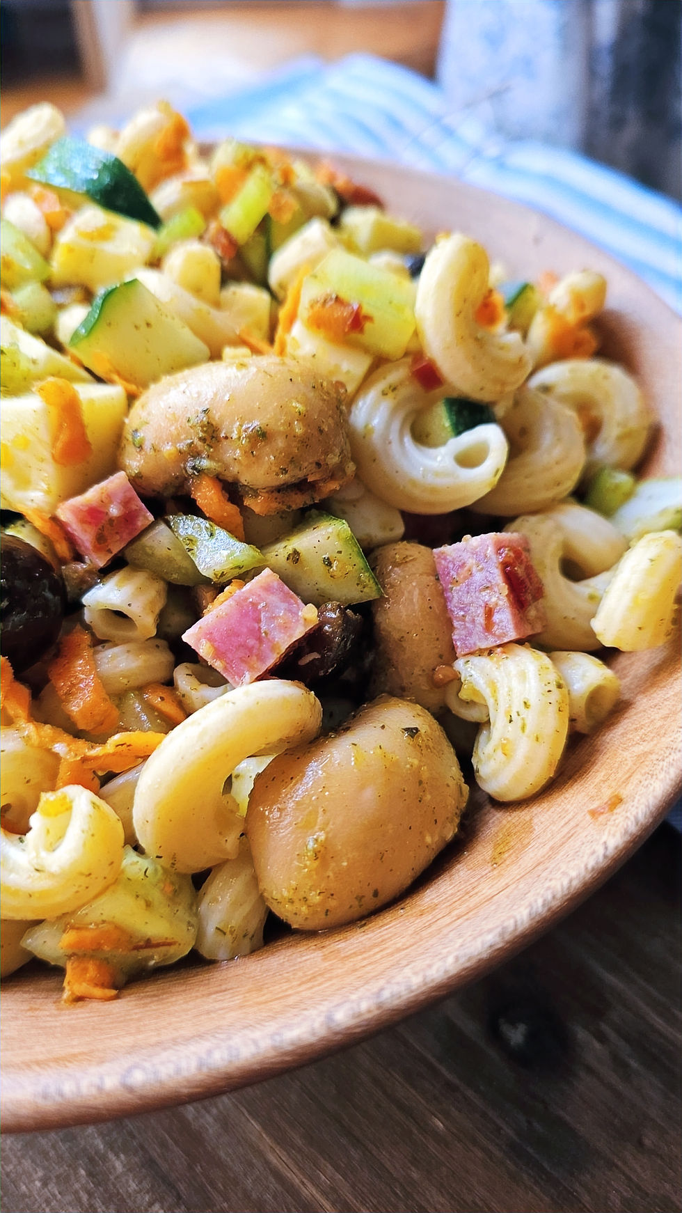 Macaroni salad with zucchini, ham, mushrooms, and carrots in a wooden bowl on a wooden surface. Bright colors create a fresh, vibrant look.