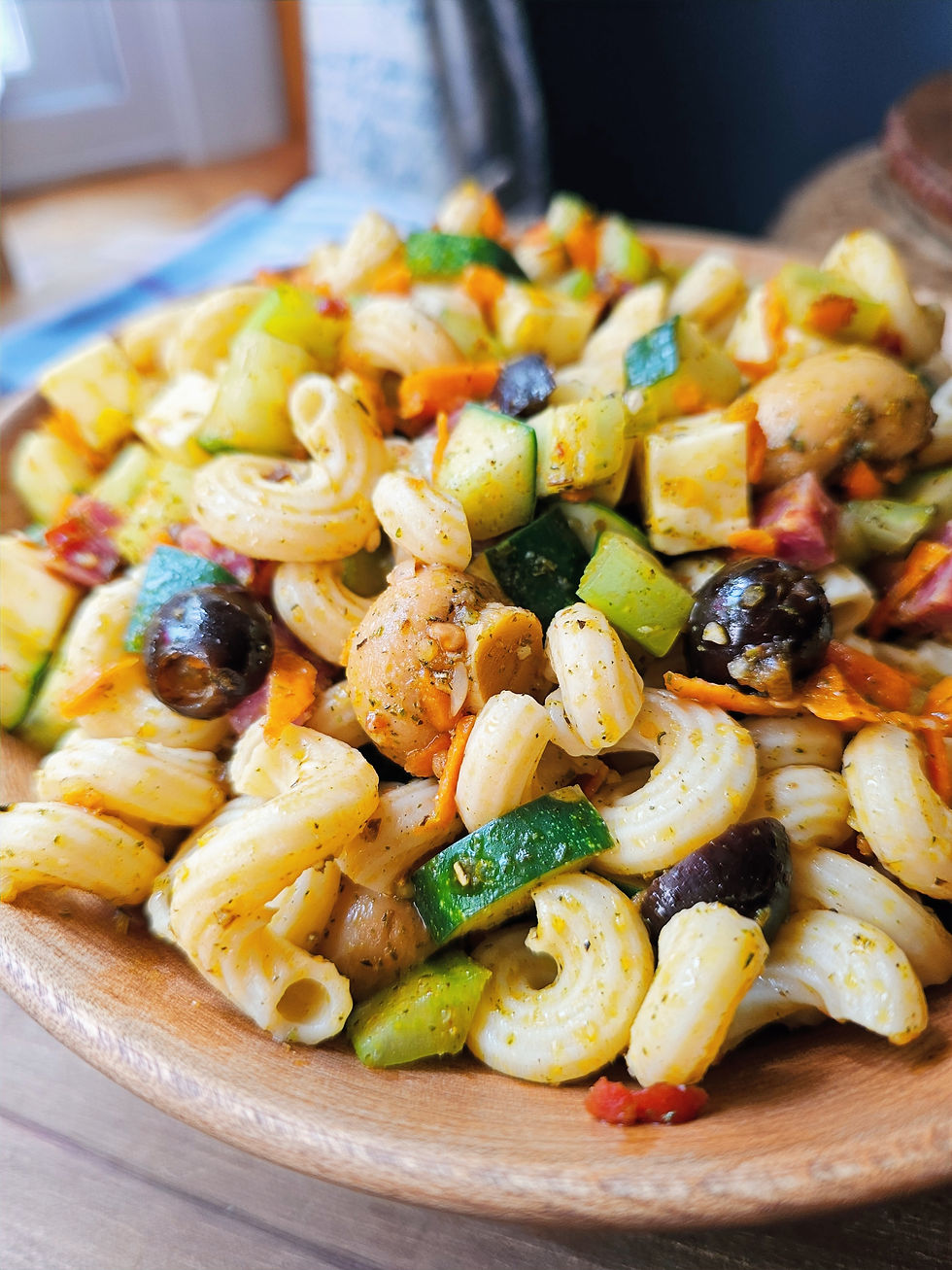Colorful pasta salad with zucchini, olives, carrots, and herbs in a wooden bowl on a table. Bright, appetizing, and fresh setting.