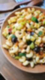 Wooden bowl with colorful pasta salad featuring olives, zucchini, and carrots, set on wooden table with wooden utensils in the background.