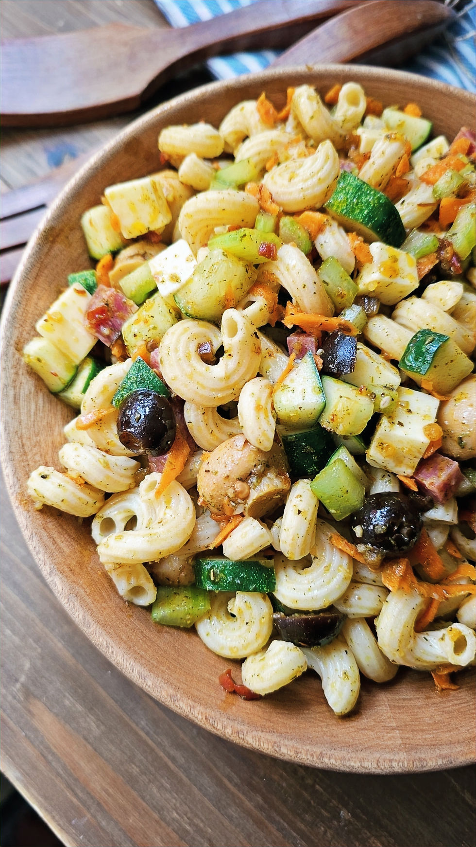 Wooden bowl with colorful pasta salad featuring olives, zucchini, and carrots, set on wooden table with wooden utensils in the background.