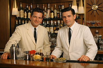 Smiling bartender in a white jacket holds a glass, standing behind a bar with bottles and glasses. Vintage black and white setting.