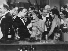 Elegant party scene with guests in formal attire holding drinks, smiling and conversing at a bar. Crystal glasses and decanters in view.