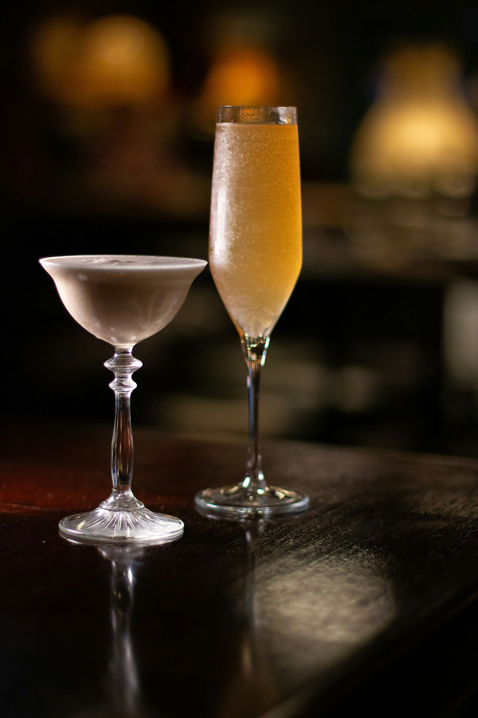 Two elegant cocktail glasses on a dark wooden bar. One glass is filled with frothy white liquid, the other with golden champagne. Dim, cozy ambiance.