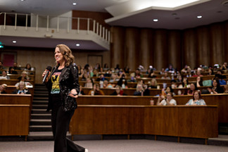 A woman holding a microphone and speaking in an auditorium.  She is wearing a black sequined jacket, black slim trousers and a retro band t-shirt.  She is smiling and engaging with the audience.