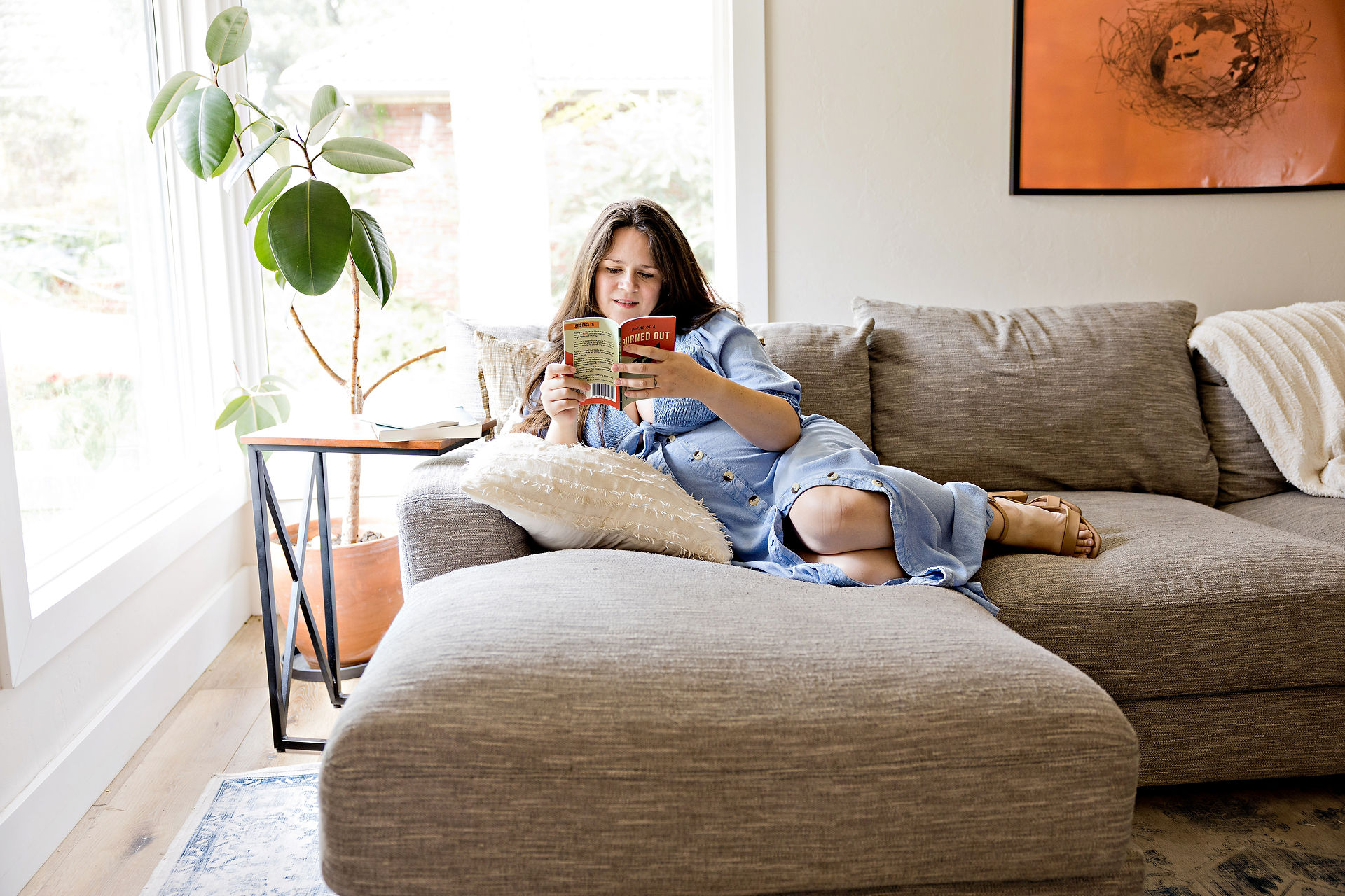 A pregnant woman wearing a blue denim dress is sitting on a sofa reading a book