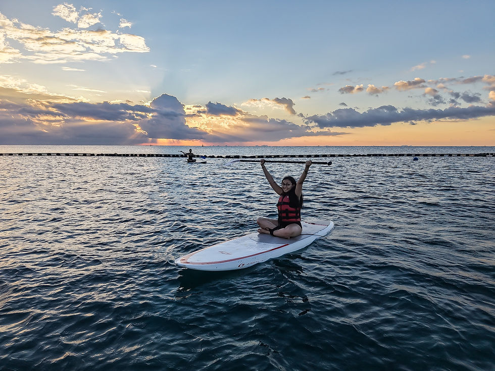 Woman on paddleboard at sunset raises arms in joy. She's wearing a red life vest. The sky has colorful clouds and a distant horizon.