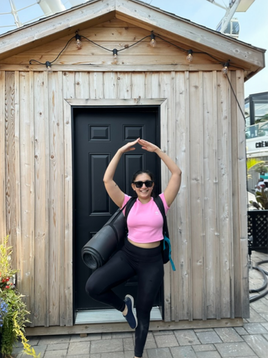 Smiling woman in a pink top doing tree pose with a yoga mat in front of a wooden hut at Old Port Montreal, Ferris wheel in the background.