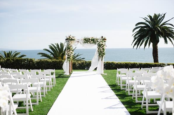 Bel-Air Bay Club lawn ceremony with white chairs lined in rows and and alter with white roses archway. An ocean view with Palm trees in the background.