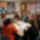 Six people sit at a table in a classroom, engaged in discussion with papers and laptops. Bulletin boards with charts and text are on walls.