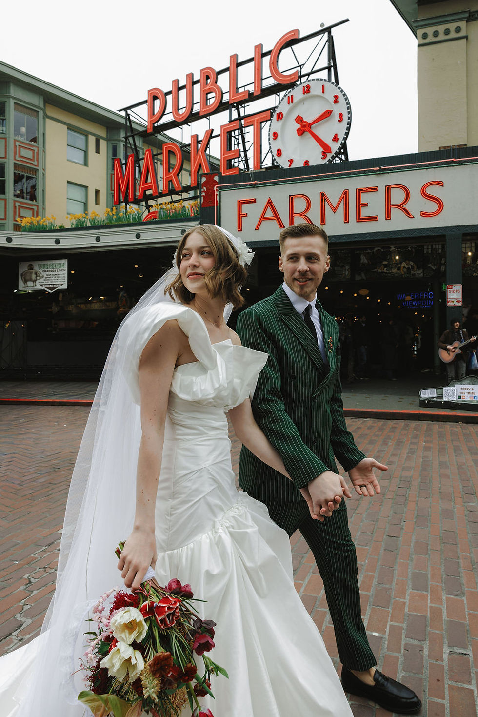 Bride in white gown and groom in green suit hold hands outside Pike Place Public Market, with neon sign and clock behind.